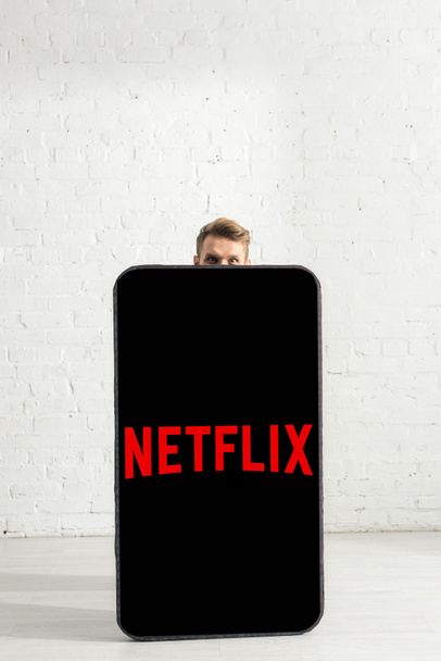 KYIV, UKRAINE - FEBRUARY 21, 2020: Man looking at camera near big model of smartphone with netflix app at home  - 写真・画像