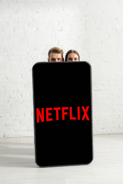 KYIV, UKRAINE - FEBRUARY 21, 2020: Couple looking at camera near huge model of smartphone with netflix app at home  - Фото, изображение