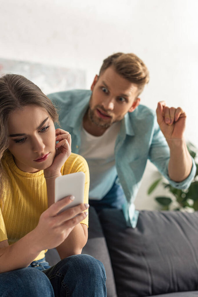 Вибірковий фокус дівчини за допомогою смартфона біля розлюченого чоловіка у вітальні
 - Фото, зображення