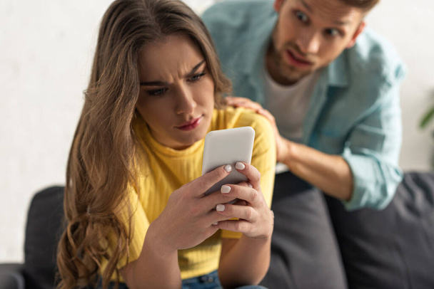 Selective focus of confused girl using smartphone near angry boyfriend in living room - Photo, Image