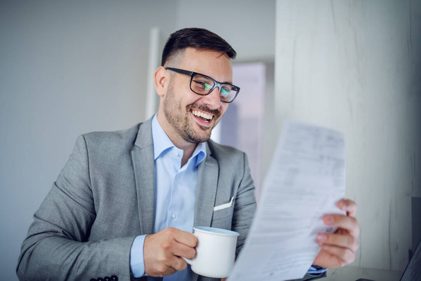 Beautiful caucasian classy smiling businessman in suit and with eyeglasses holding in one hand coffee and in other document and looking at it. On desk are laptop and paperwork. Office interior. - Photo, Image