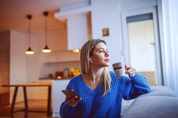 Profil pięknej, zamyślonej białej blondynki pijącej kawę i korzystającej ze smartfona siedząc w salonie na sofie i patrząc przez okno. W uszach są słuchawki niebieskozębne. - Zdjęcie, obraz