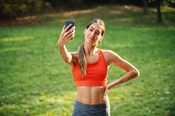 Genç gülümseyen çekici sporcu kadın ayakta duruyor ve doğada selfie çekiyor.. - Fotoğraf, Görsel