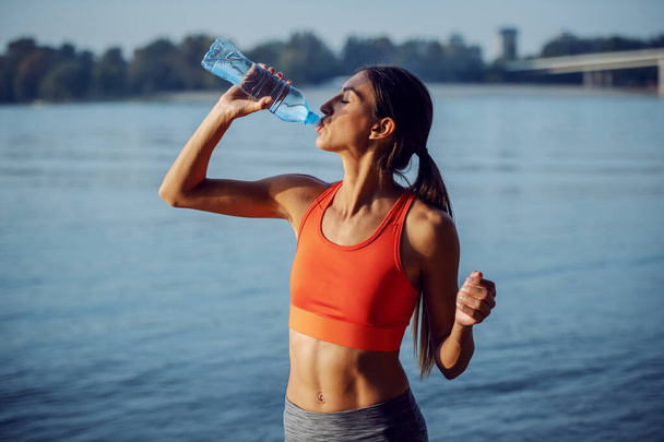 Молодая привлекательная спортсменка, испытывающая жажду, отдыхает после занятий спортом и пьет пресную воду. На заднем плане река
. - Фото, изображение