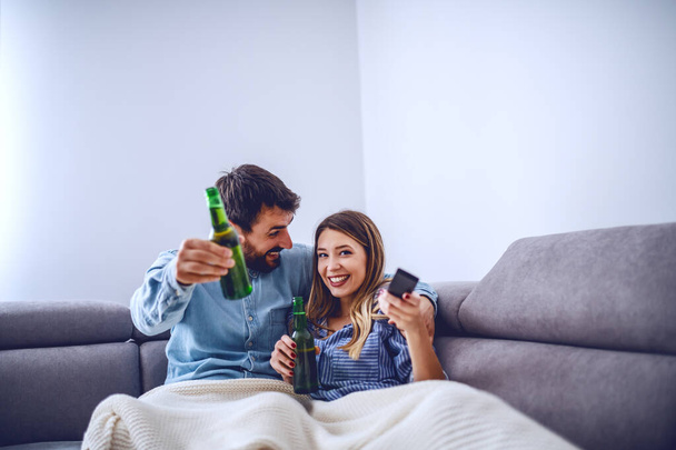 Atractiva pareja caucásica cubierta de manta sentada en la sala de estar, viendo la televisión y bebiendo cerveza. Mujer sosteniendo control remoto y cambiando de canal
. - Foto, imagen