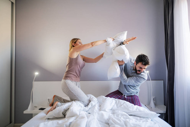 Νεαρό παιχνιδιάρικο ζευγάρι έχει μαξιλαροπόλεμο το πρωί στην κρεβατοκάμαρα. - Φωτογραφία, εικόνα