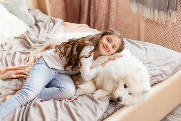 Χαμογελώντας παιχνιδιάρικο χαριτωμένο κοριτσάκι αγκαλιάζει μεγάλο λευκό χνουδωτό σκυλί Samoyed στο σπίτι, στην κρεβατοκάμαρα στο κρεβάτι - Φωτογραφία, εικόνα