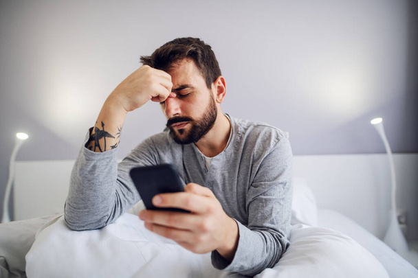 Молодой красивый бородатый мужчина сидит в спальне, держит смартфон и нервничает из-за сообщения, которое он получил от своей девушки
. - Фото, изображение