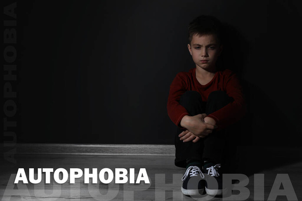 Sad little boy sitting alone near black wall, space for text. Autophobia - Фото, изображение