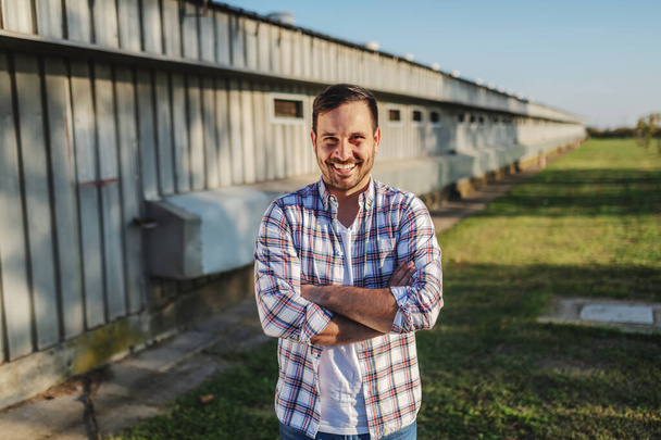 Hermoso granjero caucásico sonriente en camisa a cuadros y jeans de pie al aire libre con los brazos cruzados. En el fondo es granero
. - Foto, Imagen
