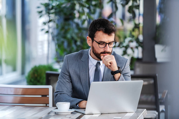 Töprengő összpontosított fehér szakállas üzletember öltönyben szemüveggel ül a teraszon a kávézó és a laptop. - Fotó, kép