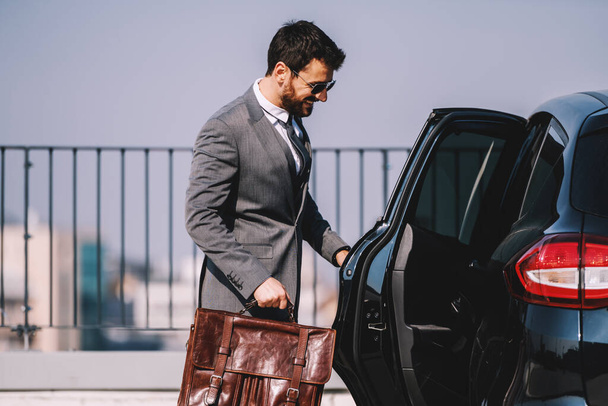 Joven hombre de negocios caucásico barbudo sonriente con traje que sostiene el maletín y entra en su coche
. - Foto, imagen