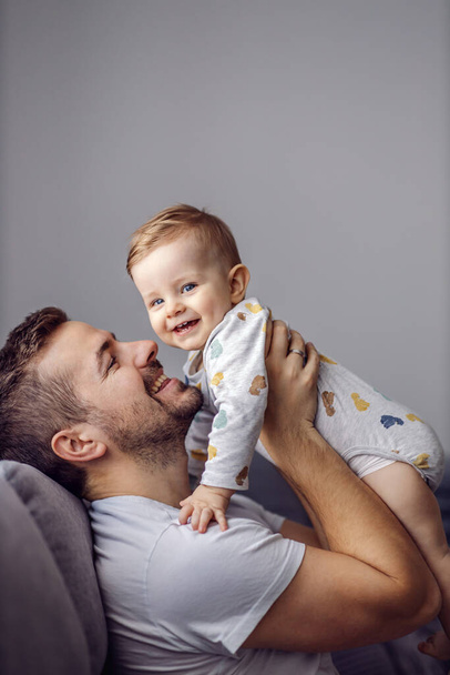 Der junge gutaussehende Vater hält seinen geliebten kleinen Sohn und spielt mit ihm, während er im Wohnzimmer sitzt. Kleinkind blickt in Kamera und lächelt. - Foto, Bild