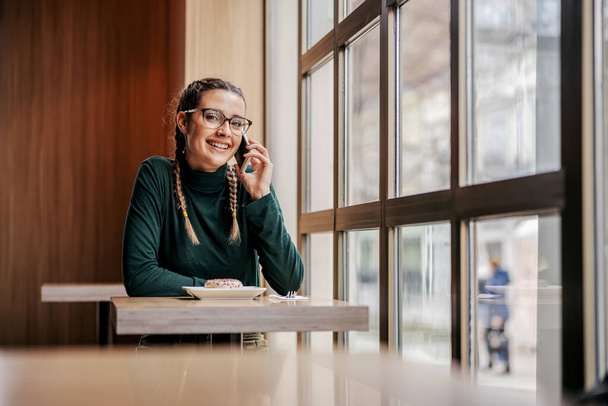 Mulher sorridente sentada na cafetaria ao lado da janela e conversando por telefone. Conceito global de telecomunicações
. - Foto, Imagem