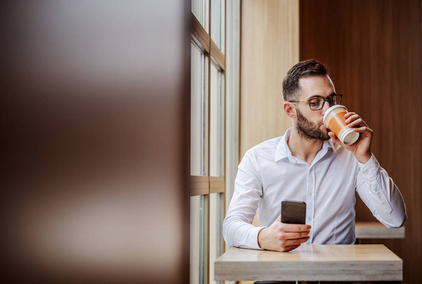 Fiatal stréber férfi elegánsan öltözött ül cafeteria mellett ablak, iszik kávét eldobható csésze és használ okos telefon ellenőrzésére üzeneteket a közösségi média.. - Fotó, kép