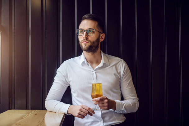 Young thoughtful geeky businessman leaning on the table next to window and looking trough it, holding glass of beer and relaxing after work. - Фото, изображение