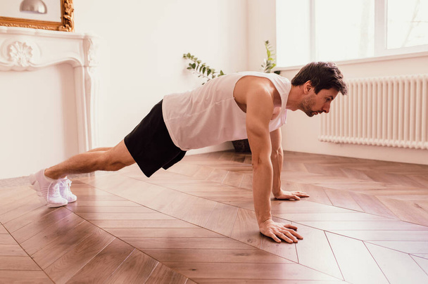  Handsome muscular man in a t-shirt and shorts doing functional exercises on floor at home - Фото, зображення