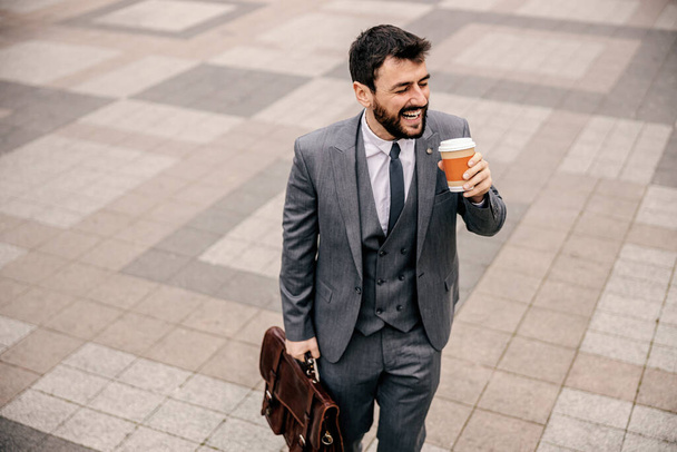 Jonge glimlachende modieuze zakenman die 's morgens koffie drinkt uit wegwerpbeker en naar zijn werk gaat. - Foto, afbeelding