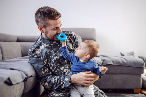 Atraktivní vousatý voják sedící na podlaze v obývacím pokoji, držící svého milovaného roztomilého chlapečka, držícího v ústech uzdu a škádlícího ho. - Fotografie, Obrázek