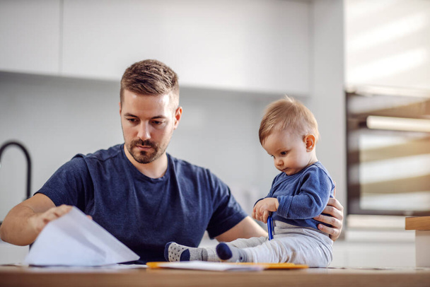 Young attractive bearded father sitting at dinning table with his beloved adorable son and paying bills online. His boy holding pen and trying to help him. - Photo, Image