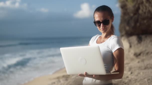 Γυναίκα ελεύθερος επαγγελματίας σε γυαλιά ηλίου κατέχει φορητό υπολογιστή, τους τύπους και τις εργασίες στην τροπική παραλία - Πλάνα, βίντεο