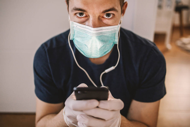 Άντρας με μάσκα και γάντια ακούει μουσική και πληκτρολογεί στο smartphone. Μείνε σπίτι. Παγκόσμια πανδημία, coronavirus. - Φωτογραφία, εικόνα