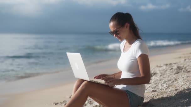 Γυναίκα σε γυαλιά ηλίου λειτουργεί στην παραλία με φορητό υπολογιστή, ανοίγει και αρχίζει να πληκτρολογεί - Πλάνα, βίντεο