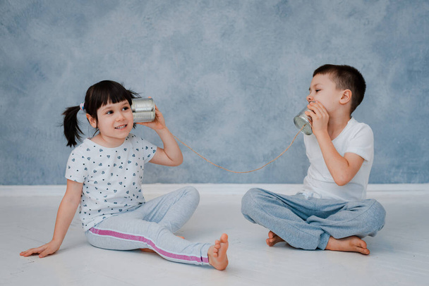 Młody brat i siostra rozmawiają z blaszanym telefonem pod szarą ścianą. Koncepcja komunikacji dla dzieci. - Zdjęcie, obraz