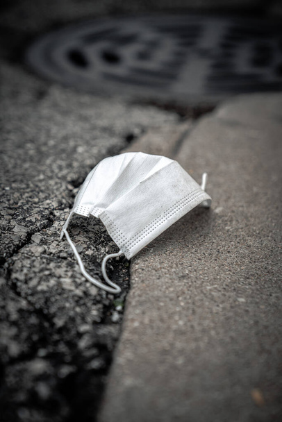 Gros plan d'un masque médical blanc sale usagé ou d'un masque respiratoire chirurgical jeté et déposé comme une poubelle sur une rue asphaltée à côté d'une bordure en béton et d'un trou d'égout pluvial dans la ville
. - Photo, image