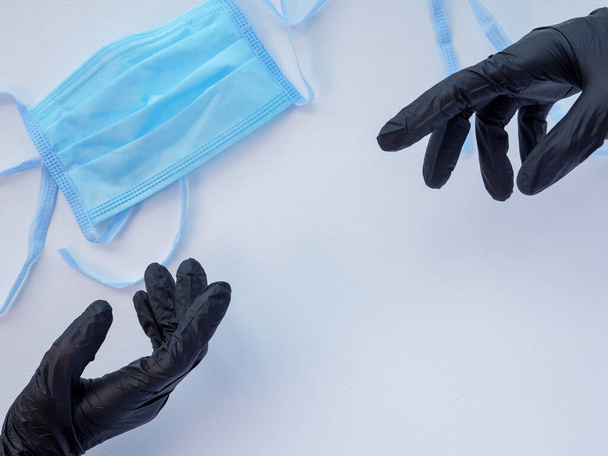 Twee handen in zwarte latex handschoenen die naar elkaar reiken. Blauw beschermend medisch gezichtsmasker op een witte achtergrond. Antivirale bescherming tijdens coronaviruspandemie. Ruimte voor tekst. - Foto, afbeelding