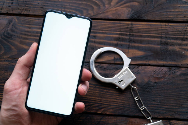 Рука мужчины держит современный смартфон с пустым экраном для текста или логотипа, наручники лежат на темном деревянном столе. Концепция технологической зависимости
 - Фото, изображение