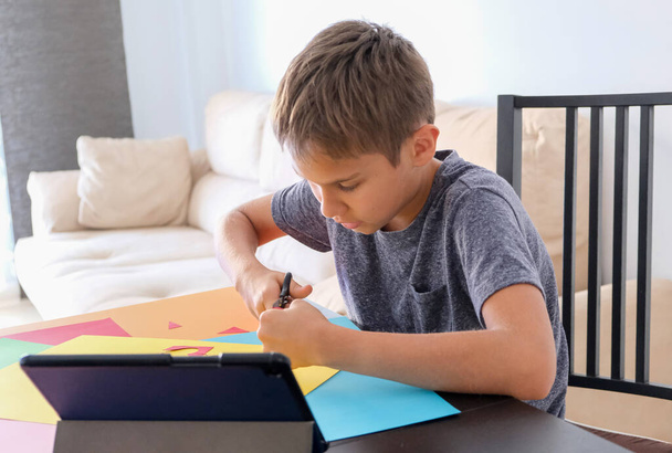 Oktatás, papírmunka gyerekeknek. A gyermek ollóval vágja a színes papírt. Fiú keres video tutorial és hogy papír kézműves projekt otthon - Fotó, kép