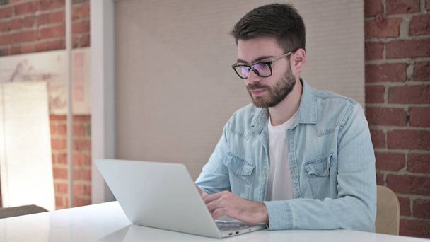 Сосредоточенный молодой человек в очках, работающий над ноутбуком
 - Фото, изображение