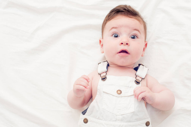 cute baby in white overalls lying on a white sheet. He is looking at the camera with a cute expression. - Photo, Image