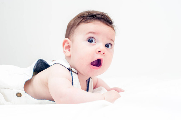 mooie baby met een leuke uitdrukking in witte overall liggend op zijn buik in een wit laken en een witte achtergrond. - Foto, afbeelding