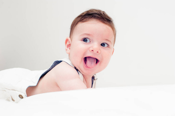 schattig baby glimlachen met zijn tong uit in witte overall liggend op zijn buik in een wit laken. - Foto, afbeelding