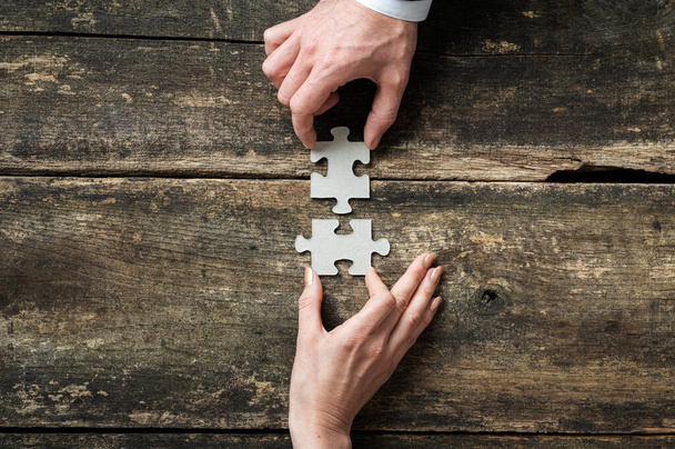 Horní pohled na mužskou a ženskou ruku spojující dva odpovídající puzzle kousky přes rustikální dřevěné pozadí. - Fotografie, Obrázek