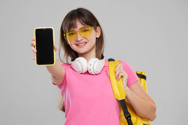 A young Asian girl in heart-shaped glasses holds a mobile phone in her hand in front of her on a pink background. New Gadget Advertising - Foto, Imagem