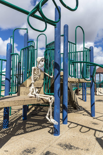 Skelet zittend op kinderspeelgoed in een leeg gesloten park op een mooie dag - Foto, afbeelding