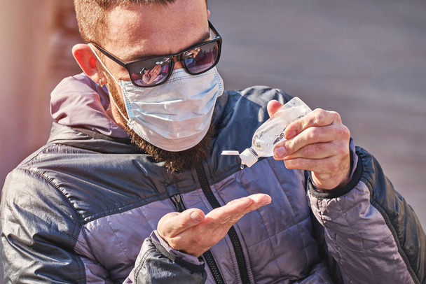 Man wearing mask uses antibacterial gel on his hands sitting in park outdoor. Self precautions during pandemic of Coronavirus. - Photo, Image