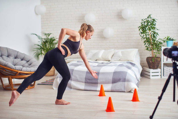 Athletic woman blogger in sportswear shoots video on camera as she does exercises at home in room. Sport and recreation concept. Healthy lifestyle. Fitness vlogger live broadcast of training session - Photo, image