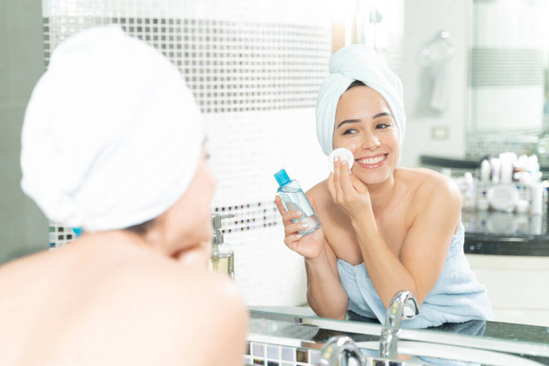 Menina bonita usando um hidratante de pele com uma almofada de algodão enquanto se olha no espelho em um banheiro
 - Foto, Imagem