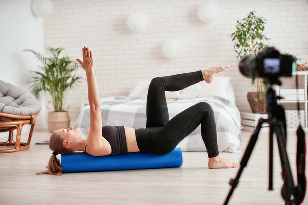 Athletic woman blogger in sportswear shoots video on camera as she does exercises at home in room. Sport and recreation concept. Healthy lifestyle. Fitness vlogger live broadcast of training session - 写真・画像