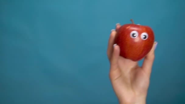 Red apple with eyes in a woman hand. Apple jumps into the frame and looks around on a blue background. Woman hand close-up. - Video