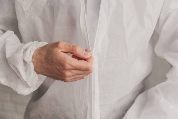 Doctor epidemiologist puts on white protective suit, fastens a zipper, hands close-up. Hands in protective rubber gloves. Personal protective equipment concept. - Photo, image