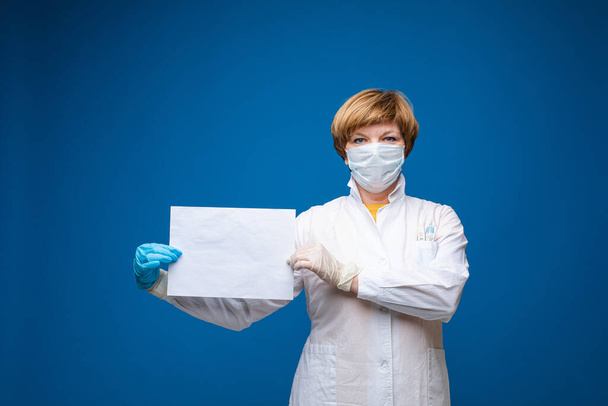 Beautiful caucasian woman with medical mask holds a white sheet of paper, portrait isolated on blue background - Fotó, kép