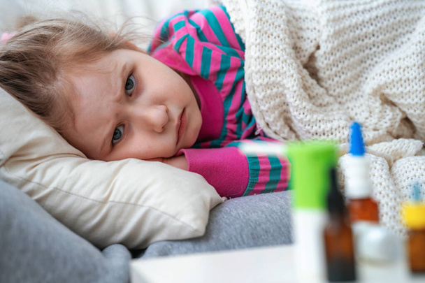 Sick little girl is lying on sofa, wrapped in a blanket. on the table are medicines. An unhealthy sad child is being treated for a viral disease, a cough medicine. epidemic of coronavirus, kovid-19. - Foto, Imagem