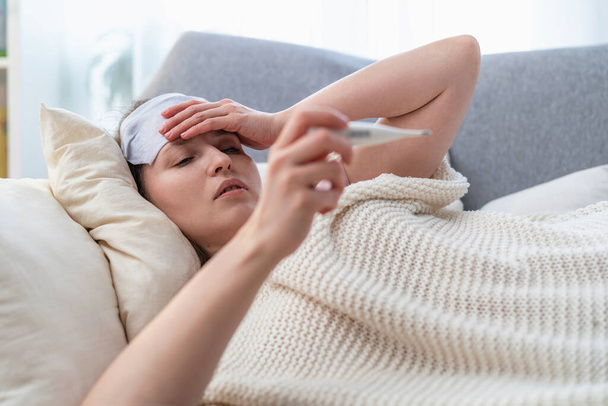 Portrait of a young sick girl lying on sofa with her hand on her head. A young woman has high temperature, takes her temperature and coughs. Concept of treatment viral diseases. Coronavirus, covid-19 - Foto, Imagen