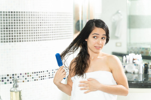 Jolie femme enveloppée dans une serviette et brossant ses cheveux emmêlés dans la salle de bain
 - Photo, image