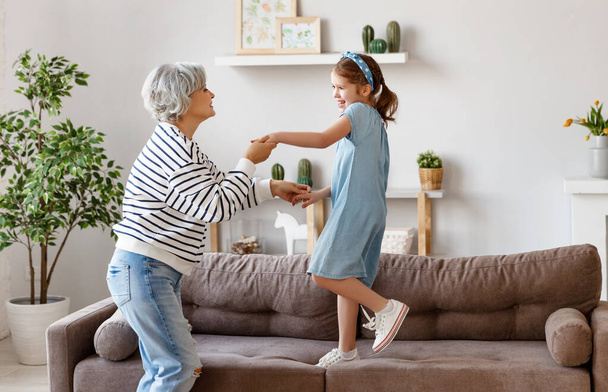 Seitenansicht einer grauhaarigen Frau in gestreiftem Hemd und Jeans, die mit ihrer fröhlichen Enkelin im gemütlichen Wohnzimmer tanzt, während sie die Zeit zusammen bei hom verbringt - Foto, Bild